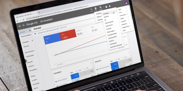 A laptop displaying the Google Ads all campaigns overview with performance metrics and a dropdown menu open, representing the article's walkthrough of Performance Max campaign structure - from strategy and budget settings to asset groups, signals, and auction statistics