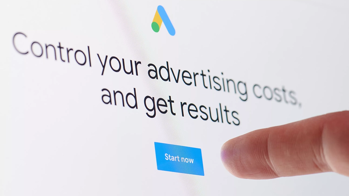 Finger pressing the "Start now" button on the Google Ads homepage with "Control your advertising costs and get results" headline