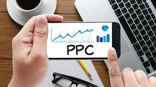 Hands holding a phone displaying PPC growth charts on a desk with a laptop, representing B2B PPC strategy management