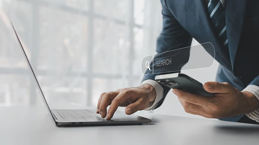 Businessman typing on a laptop while holding a smartphone with a search bar overlay, showing cross-device search behavior