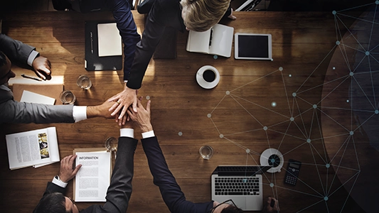 Business team stacking hands together over a table with documents and a laptop, representing partnership and joint campaign strategy
